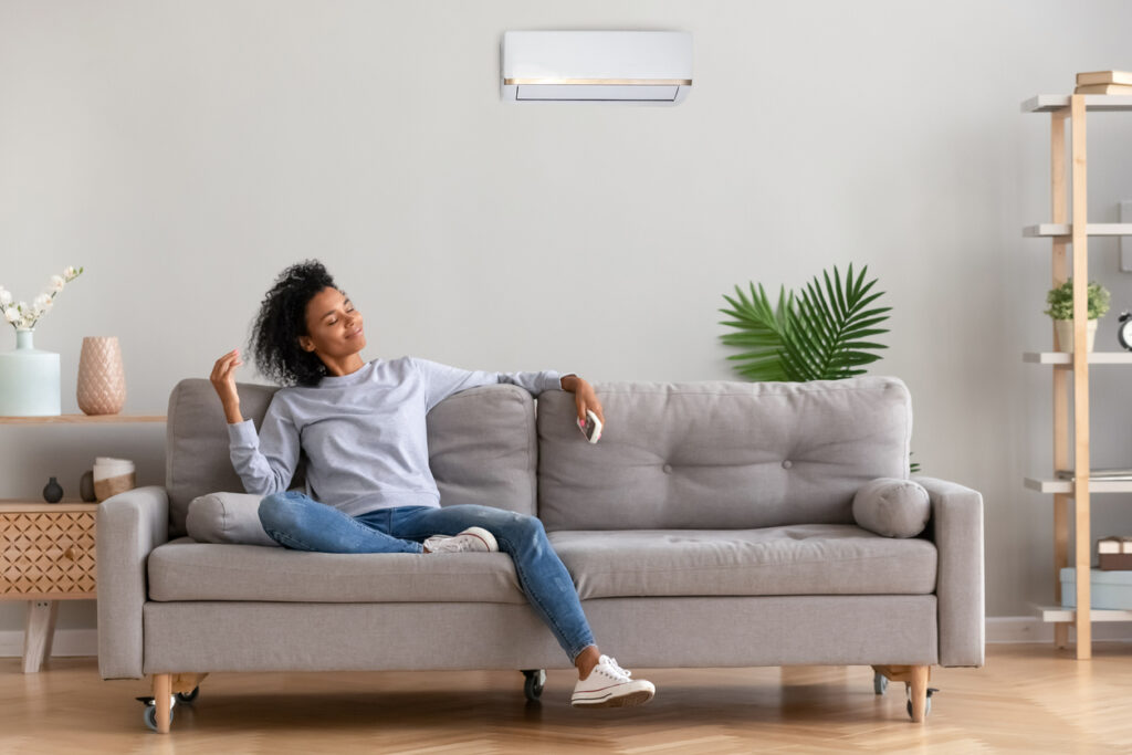 Woman sitting on light gray couch in a living room, enjoying cool air from the ductless mini-split system on the wall behind her.